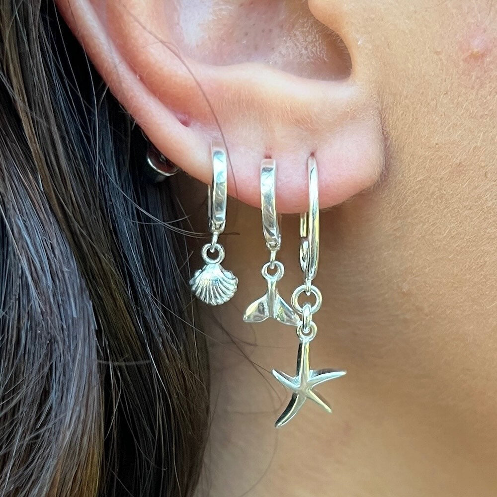 Close-up of an ear wearing silver hoop earrings with shell, mermaid tail, and starfish charms.