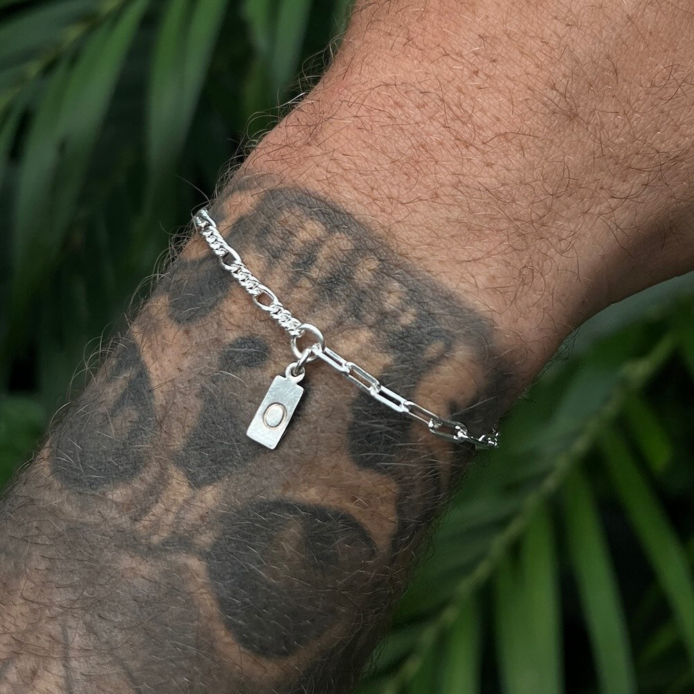 Silver chain bracelet with a tag on a tattooed arm against a green leafy background