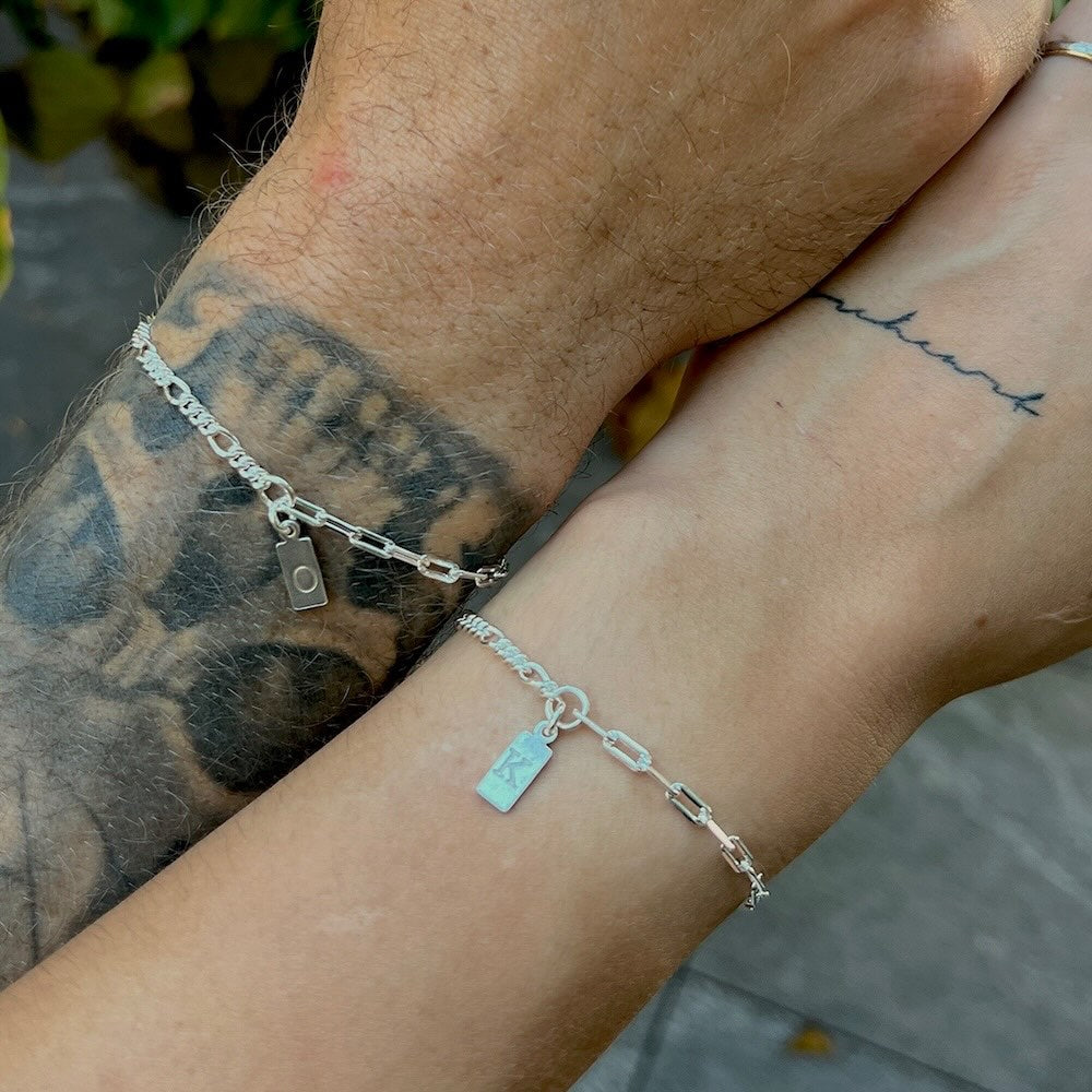 Two wrists with silver bracelets featuring small tags, one with tattoos.