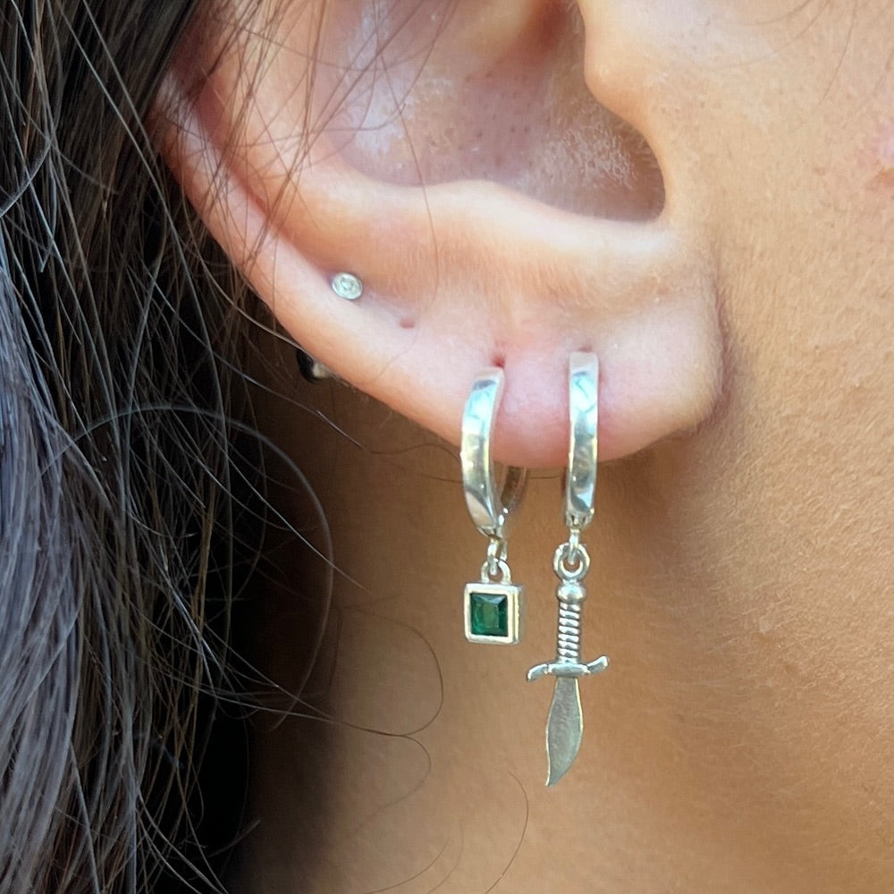 Close-up of an ear wearing silver hoop earrings with sword and green stone charms.