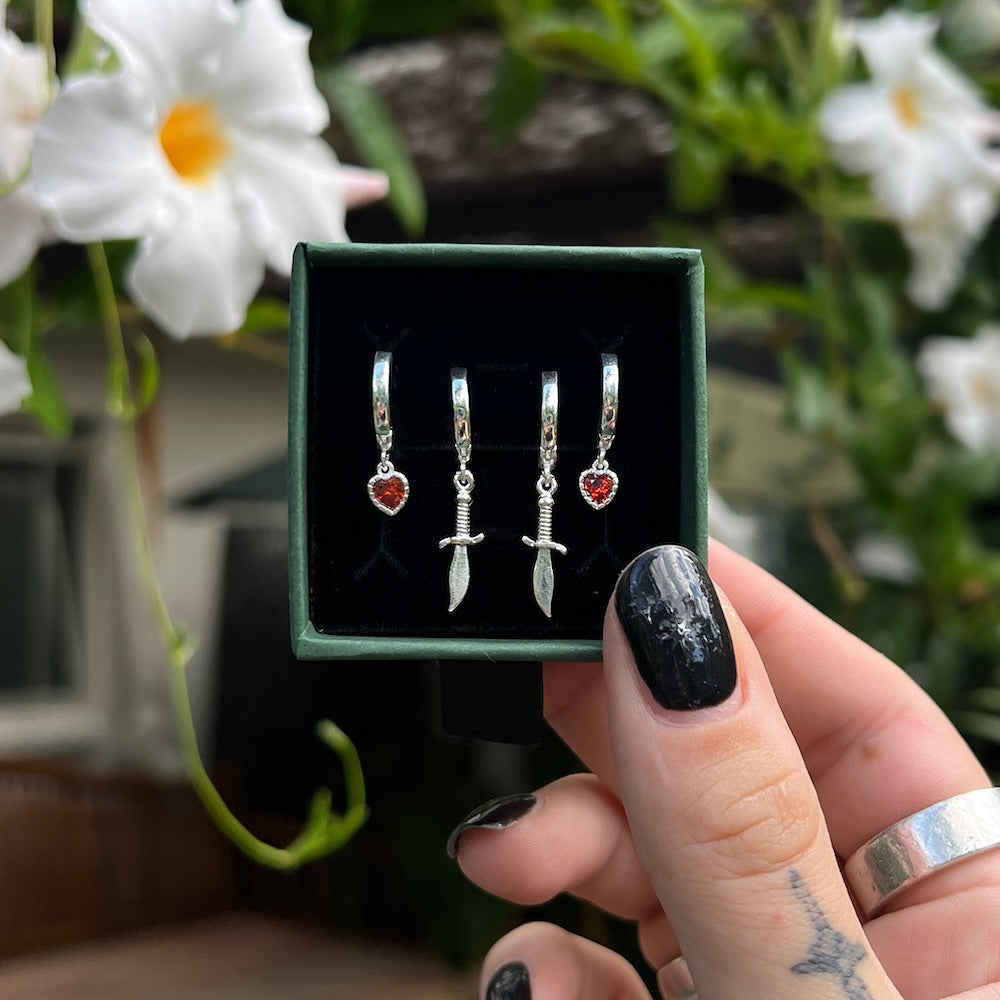 Set of silver earrings with red stones in a green box held by a hand with black nail polish.