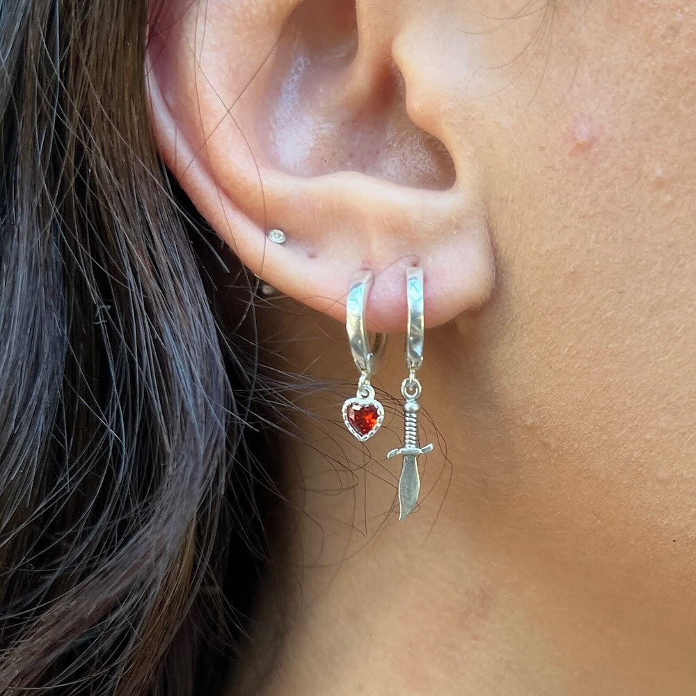Close-up of an ear wearing silver hoop earrings with heart and sword charms.