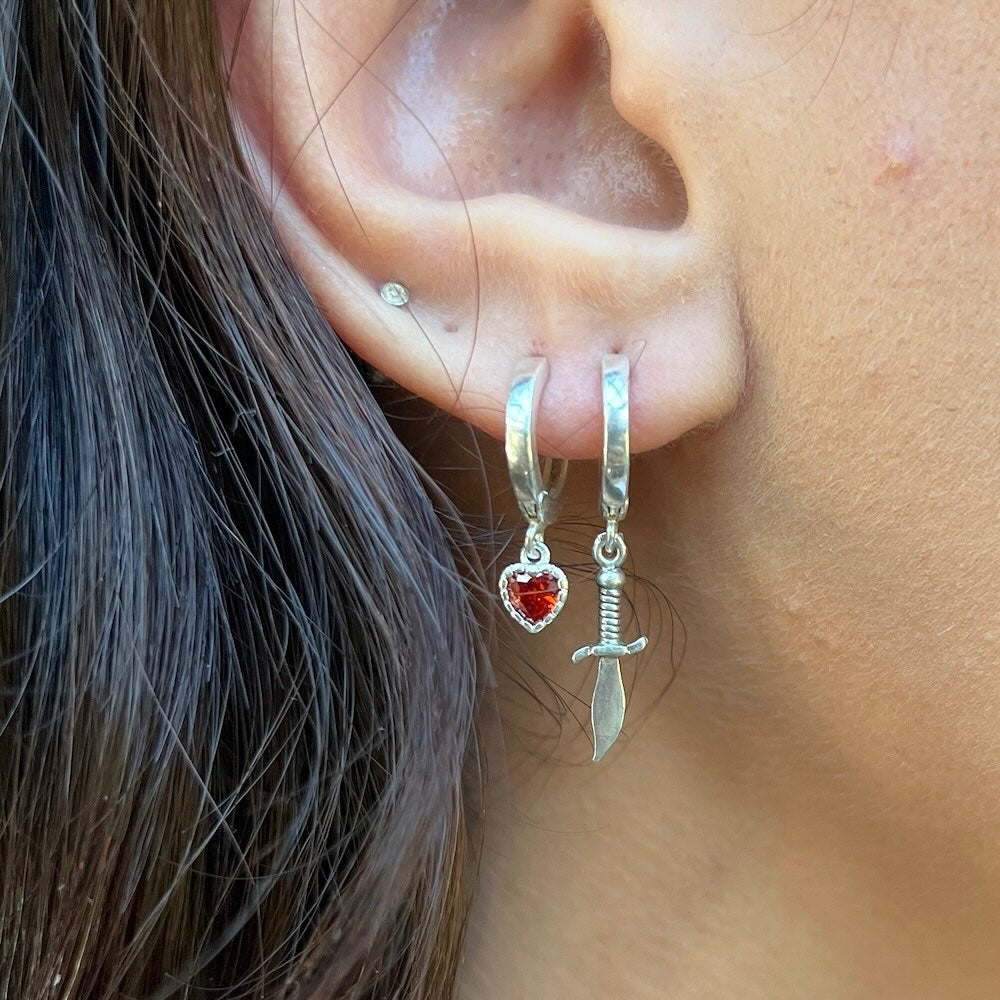 Close-up of an ear wearing silver hoop earrings with heart and sword charms.
