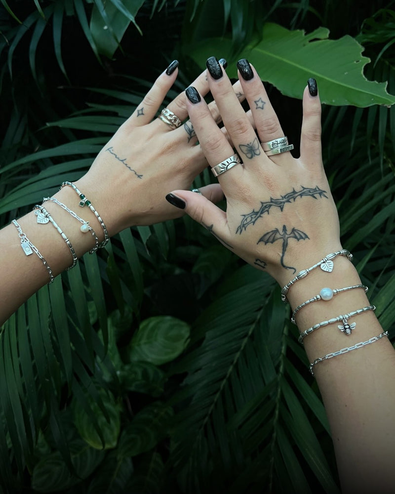 Hands with tattoos and multiple silver bracelets against a green leafy background